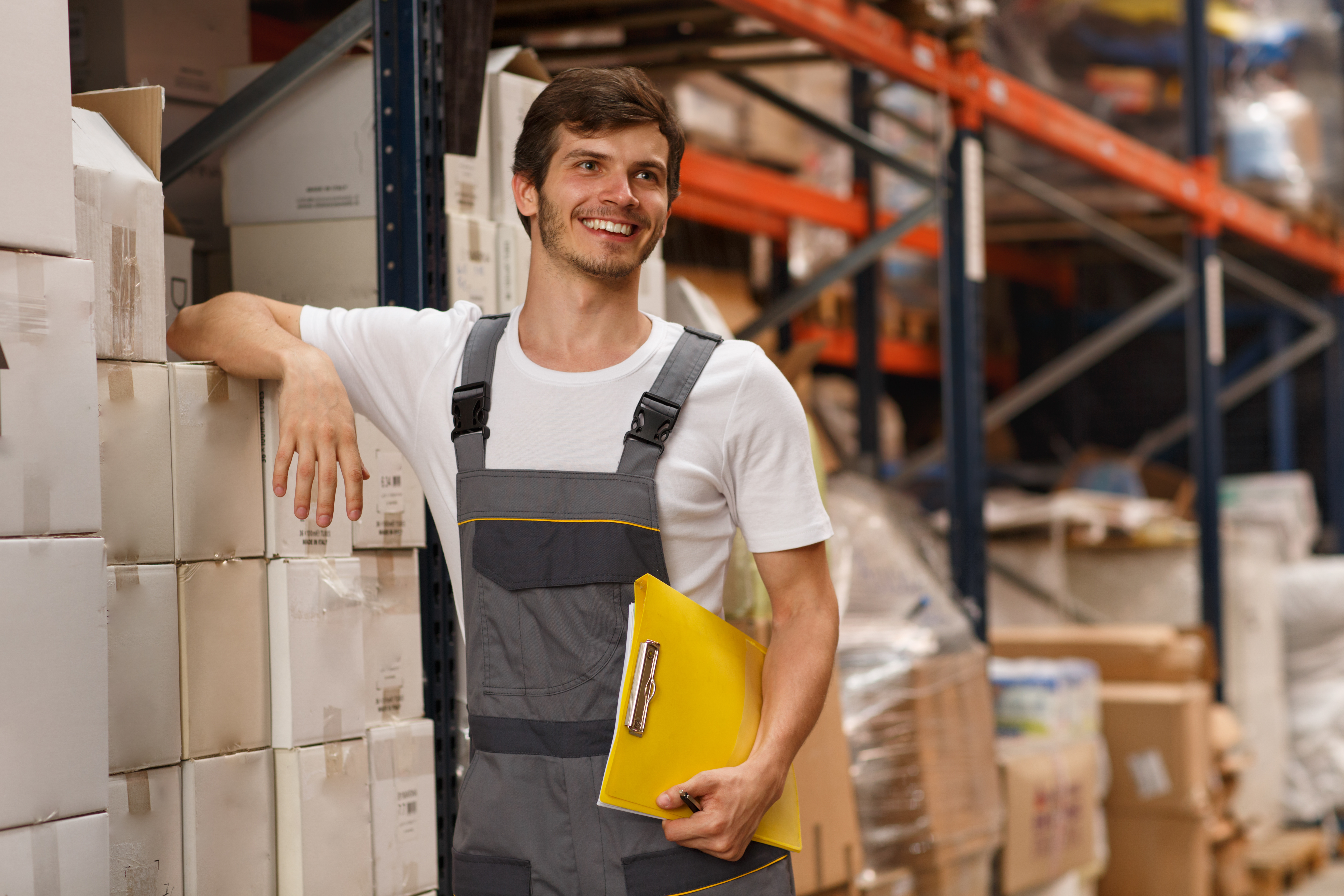 Cheerful worker wearing uniform and white t shirt, holding yellow clipboard. Handsome man smiling, standing and leaning on white boxes in warehouse. Concept of entrepot and commercial industry.